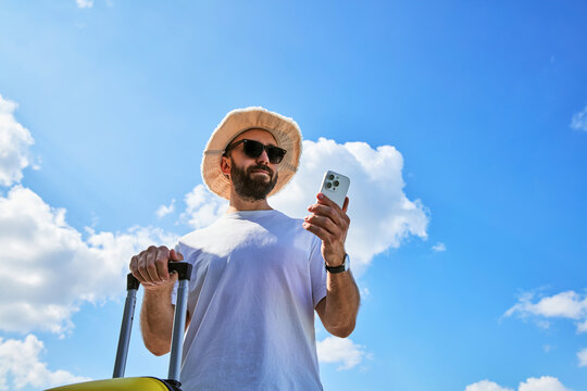 Low angle view of male traveler with suitcase checking smartphone against blue sky