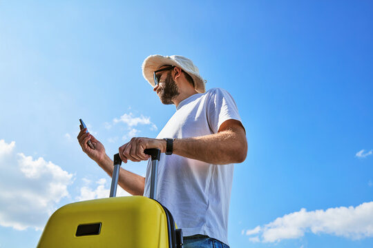 Man Traveler with Yellow Suitcase Using Smartphone Outdoors Against Blue Sky