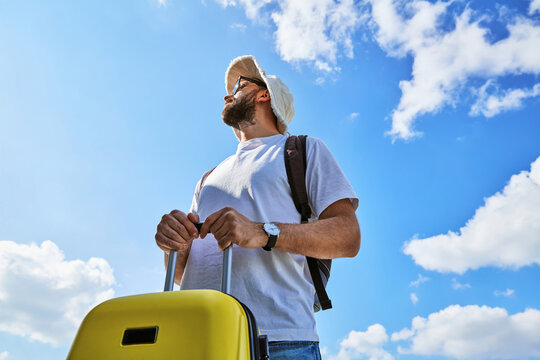 Male traveler with yellow suitcase and backpack looking up against blue sky
