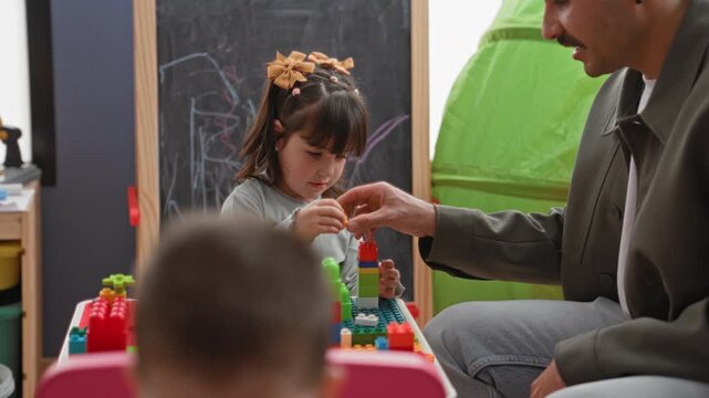 Girl stacking plastic blocks with hands while man helps beside her at the play table, chalkboard and tent visible in building; concentration learning.