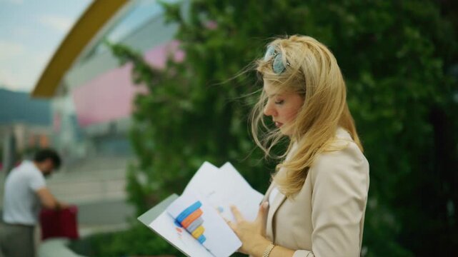 A young businesswoman is intently reviewing financial documents with charts and graphs outdoors. She meticulously examines data in her folder amidst an urban outdoor setting.