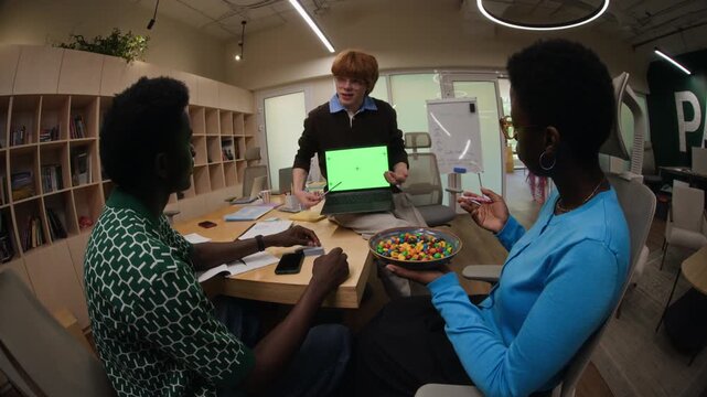 Caucasian young adult male presenting green screen laptop to African American coworkers during office meeting, camera zooming out. Clip is ideal for tech product demos and software marketing