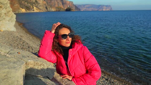 Woman beach ocean relaxes in a bright pink jacket on a stone wall by the sea during a peaceful golden hour sunset