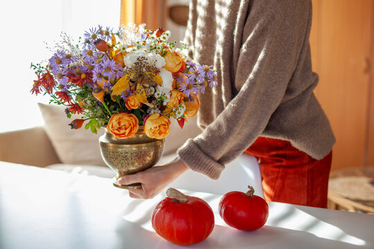 Woman decorating home for autumn with flowers arrangement, pumpkins on table in living room. Cozy Thanksgiving season. Bouquet