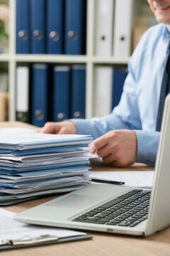 Businessman in blue shirt reviews paperwork stacked on desk with laptop and office supplies, organized filing cabinets visible in the background