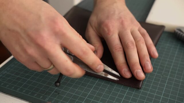 Leatherworker marking stitches on brown leather pouch with wing divider tool in a traditional workshop