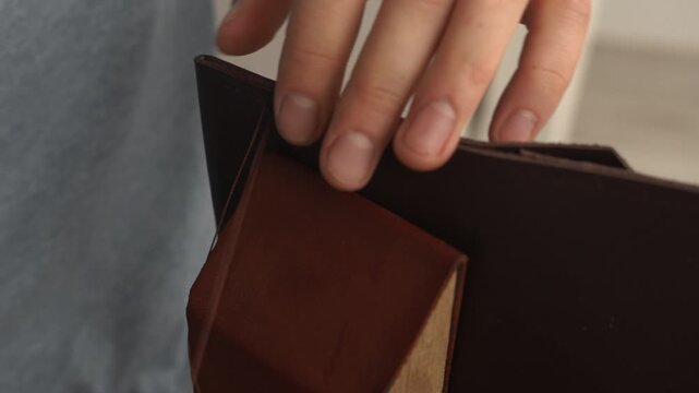 Leather craftsman hands sewing a genuine brown leather wallet with a saddle stitch using a needle and thread in a workshop