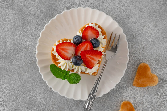 Valentines day dessert. Heart shaped cake millefeuille with vanilla cream and fresh berries on a white plate on a gray concrete background. Romantic food concept. Top view.