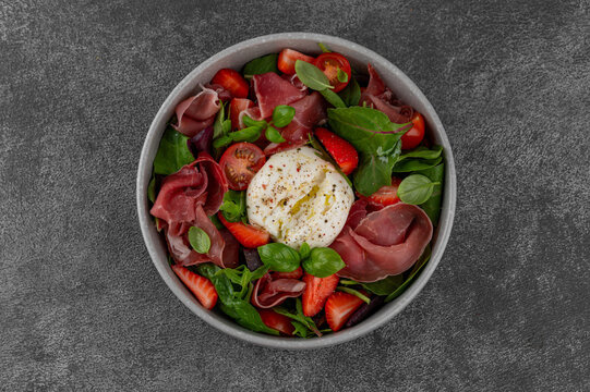 Salad with strawberries, tomatoes, burrata and jamon. Fresh summer salad in a bowl on a dark background. Top view.