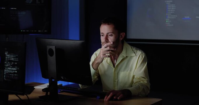 Adult Caucasian man works at computer workstation, reviews monitor and touches face while code appears on projected screen behind, supporting software analysis, digital workflow, technology planning.