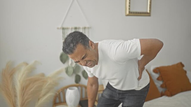 Man clutching lower back on bed inside a building, grimacing in pain while leaning forward; pain recovery.