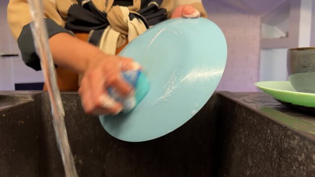 Hands washing light blue bowl under running water in kitchen sink, low-angle closeup showing scrubbing, rinsing, and soap foam during cleaning routine, with washing, kitchen, sink, hand detail