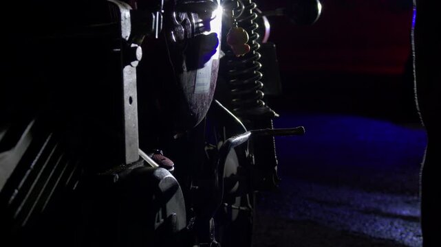 Close-up of a motorcycle being handled at night, with suspension, wheel and mechanical parts illuminated by blue light in a dark roadside setting, creating a cinematic maintenance scene