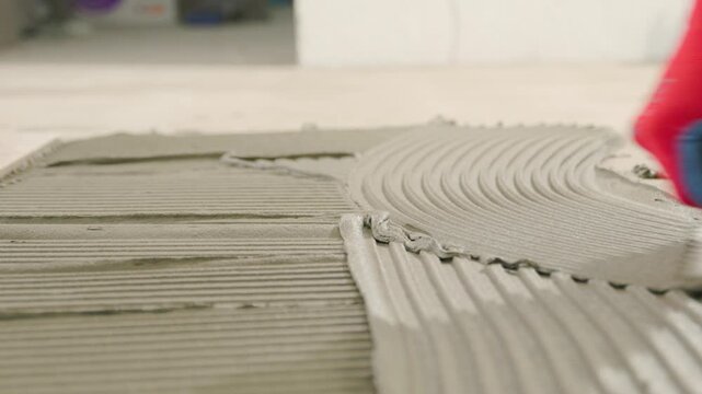 Professional tiler spreading tile adhesive with a notched trowel on the floor of a renovating house.
