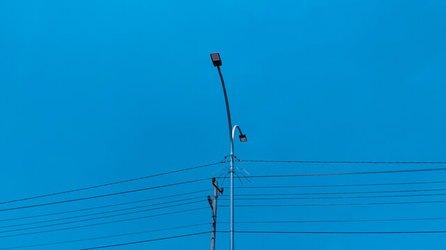 Street light pole with electric power lines and security camera against a vibrant clear blue sky background in an urban utility infrastructure setting