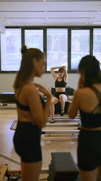 A woman performs stretching exercises on a pilates reformer machine in a bright studio. Other women observe the focused fitness session.