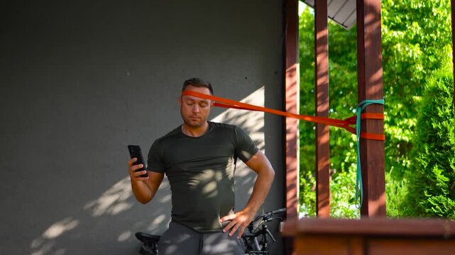 Cinematic static footage of an athletic man performing an advanced neck isometric hold using a thick heavy orange resistance band while checking his smartphone.