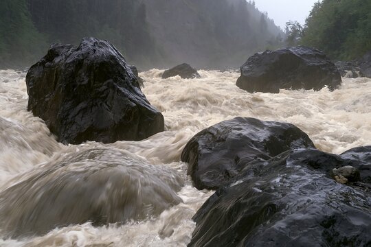 Powerful floodwater rushes around huge black boulders in a rainy mountain gorge, showing flash flood conditions, dangerous rapids and extreme rainfall