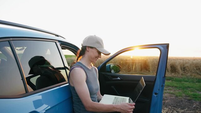 Young professional female farmer using a laptop beside a parked vehicle in the middle of a field, managing farm operations and demonstrating leadership in modern agriculture.
