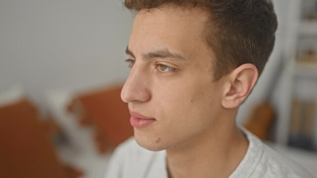 Young man face looking right, seated on bed beside orange pillows and acoustic guitar in a residential building; contemplative.