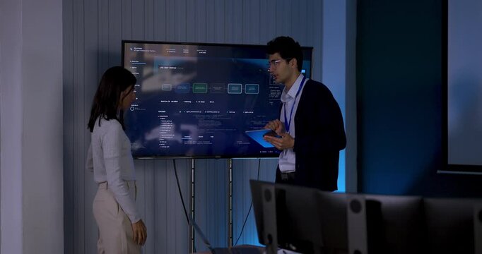 Adult male technology professional presents digital dashboard on screen, pointing at system interface while female colleague responds during collaborative software strategy meeting in modern office.
