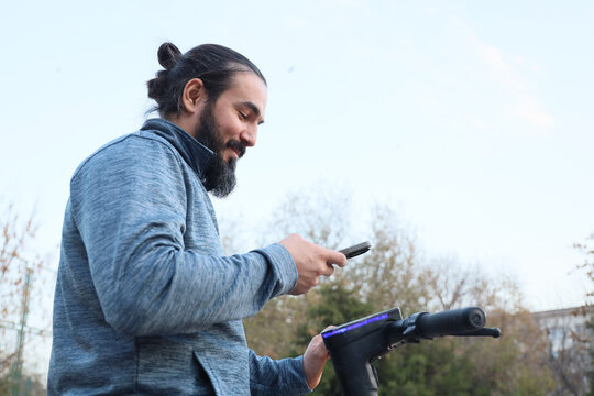 Bearded Man With Tied Back Hair Looks Down At Smartphone While Holding Electric Scooter Handle Outdoors In Daylight, Surrounded By Soft Trees And Open Sky, Suggesting Urban Mobility
