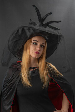 Woman wearing a witch hat with feathers and veil, black cape with red lining, and black dress, posing with an intense expression
