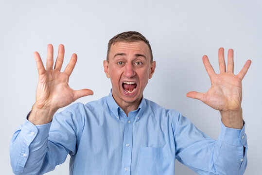 Young man wearing a blue shirt wildly screaming with eyes wide open while raising both hands, showing an expression of shock, surprise, or fright on a white background