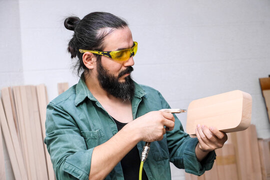 Bearded Woodworker In Yellow Safety Glasses Inspects Smooth Curved Wooden Block While Holding Pneumatic Tool Inside Workshop With Stacked Timber Boards, Focused On Careful Shaping