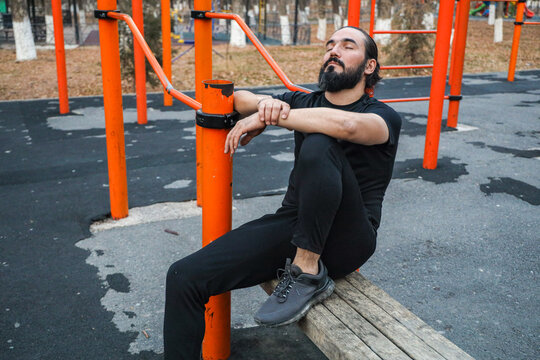 Adult Man Sits Quietly On Orange Calisthenics Equipment In Urban Park, Eyes Closed With Calm Expression, Wearing Black Sportswear And Gray Sneakers, Conveying Rest, Recovery, And Pause After