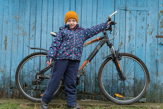 Young girl wearing a winter jacket and orange beanie, proudly standing next to her bicycle, smiling at the viewer against a weathered blue wooden fence