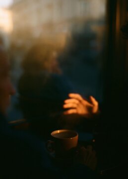 Person sitting at a table with a cup of coffee in front of them. the person is facing away from the camera, with their hands clasped together in a prayer-like gesture.