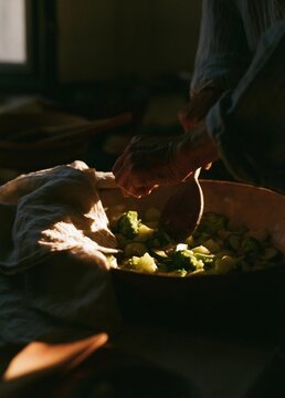 Person's hands holding a wooden spoon over a large pot of food. the pot appears to be filled with a variety of vegetables, including broccoli, cauliflower, and other leafy greens.