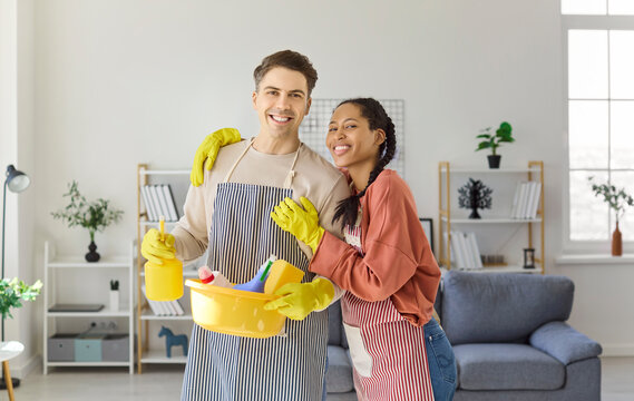 Couple man and woman cleaning with gloves teamwork. Smiling partners in aprons hold supplies and a bucket in a living room, promoting housework. Concept shows teamwork and home cleanliness.