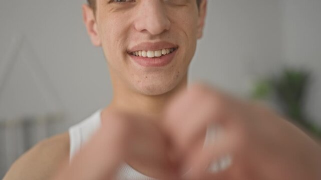 Young man smiles showing teeth, wearing white tank top, forming heart with hands framing face in studio; affection connection.