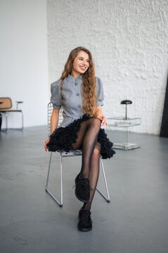 Portrait of young woman seated on metal wire chair in bright minimalist studio, smiling with eyes closed and hand touching hair, outfit includes tailored grey jacket with decorative buttons