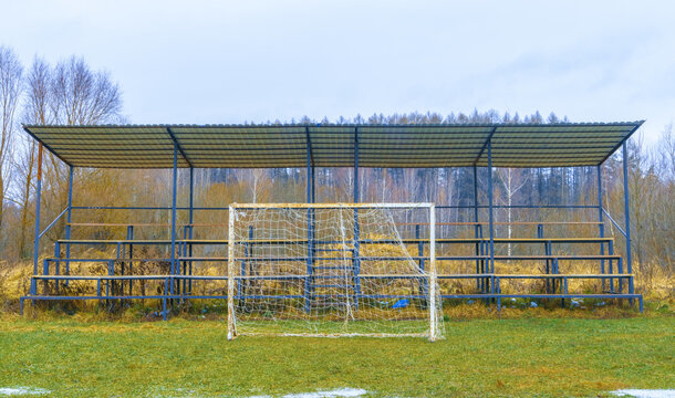Weathered metal bleachers with corrugated roof stand behind worn soccer goal positioned on overgrown grassy pitch, muted winter trees form background, soft diffuse