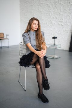 Young woman seated in modern studio wearing gray jacket and black skirt, with woman, portrait, model, sitting detail, showing chair, studio, fashion, style context