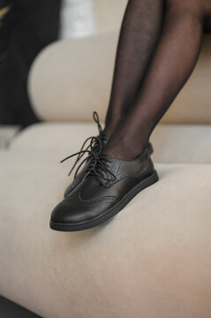Closeup of legs in sheer black hosiery and black brogues on cream sofa, with legs, hosiery, stocking, black detail, showing sheer, brogue, shoe, leather context