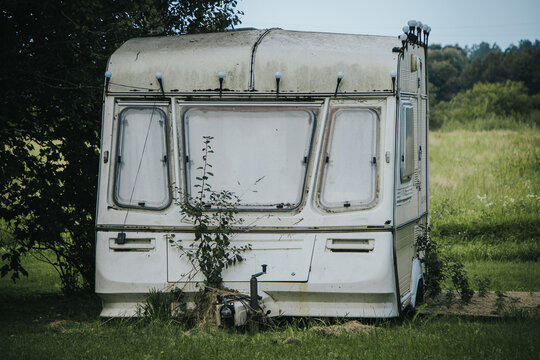 A white, weather-worn caravan standing under trees in a grassy rural field, with weeds growing around its exterior, a sign of abandonment and decay.