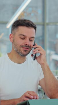Vertical video. Close up of man barista in coffee shop accepts a pre-order on a mobile phone call and writes it down on laptop computer in a cafe. Delivery food home online order concept.