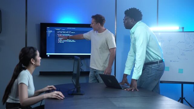 Diverse software developer team analyzing code on large monitors in dark IT office. Asian female and African American male programmers discussing technology projects. Corporate cybersecurity teamwork