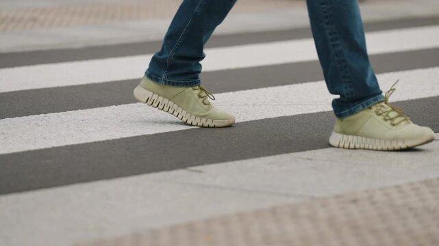 Cinematic close up view urban crosswalk with zebra stripes on asphalt, male pedestrian walking in slow motion, cropped legs to knee level, overcast cloudy city atmosphere