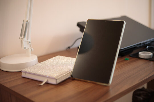 Tablet standing on wooden desk with textured notebook and desk lamp, laptop blurred in background, modern study workspace, high quality photo