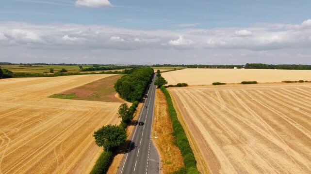 Drone view of motorcycle overtaking car on country road between harvested crop fields. Scenic rural highway surrounded by golden farmland under dramatic clouds with summer countryside landscape. Road