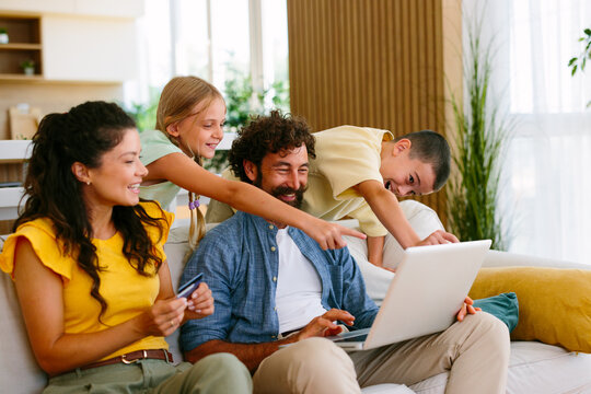 Happy family with parents and children sitting on a sofa, enjoying online shopping together on a laptop at home, preparing to make a payment with a credit card