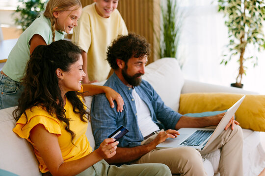 Happy family with parents and children sitting on sofa, browsing e-commerce website on laptop, and making a secure payment with a credit card for online shopping