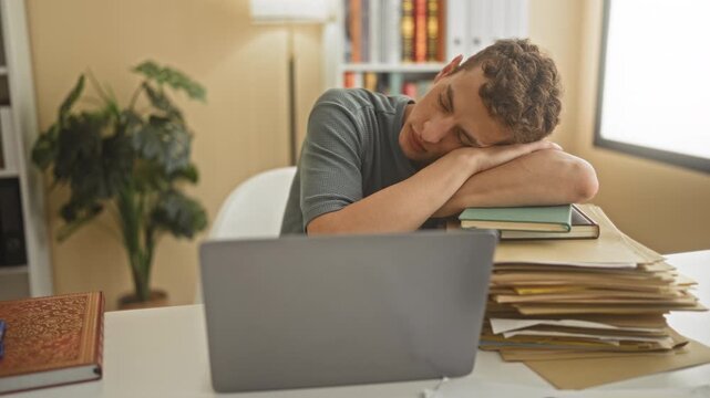 Man resting head on forearm at desk with laptop and pile of folders and books in a building; fatigue burnout.