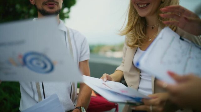 Three business people are discussing documents with charts and graphs outdoors. Colleagues are collaborating on project plans and financial data in a positive meeting.