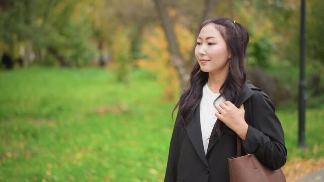 Close up of young woman with long dark hair wearing black blazer and white shirt holding lapel while standing on autumn path. Woman carries brown handbag over her shoulder as woman looks